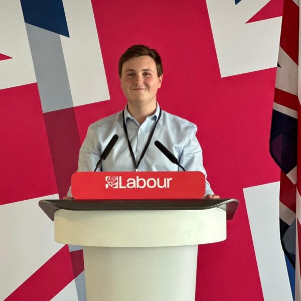 Photo of a man stood at a white podium wearing a blue shirt smiling. Podium has red sign reading "Labour". Behind the man is a backdrop with the union jack.