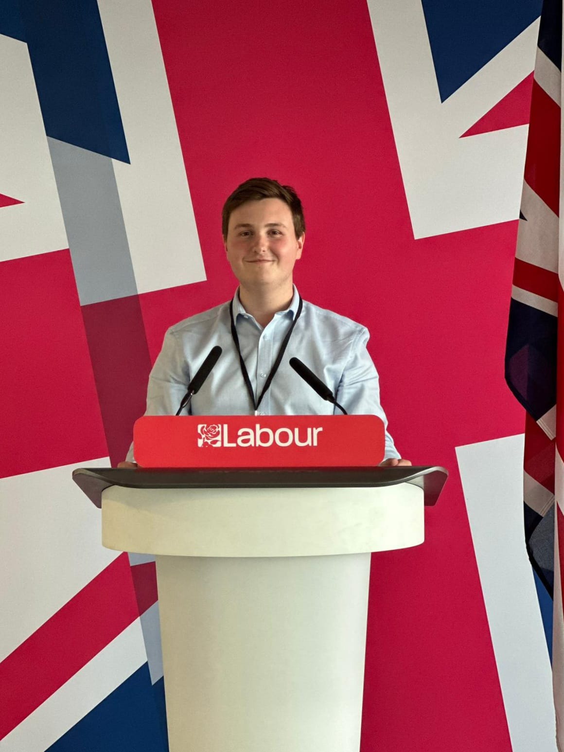 Photo of a man stood at a white podium wearing a blue shirt smiling. Podium has red sign reading "Labour". Behind the man is a backdrop with the union jack.